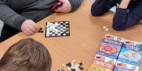 Powiększ grafikę: Przy stoliku siedzą trzej chłopcy. Na stole leżą rozłożone gry: warcaby i szachy oraz pudełka gier szachy, chińczyk, statki, inteligencja.