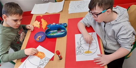Powiększ grafikę: Dwóch chłopców, jeden maluje farbammi, a drugi wylepia bibułą kontury dyni narysowanej na kartce.