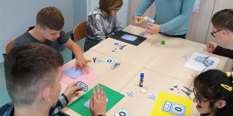 Powiększ grafikę: Pięcioro uczniów siedzi przy stole. Każdy uczeń układa robota z wyciętych elementów na kolorowych podkładkach.