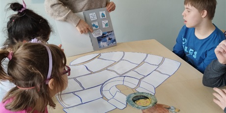 Powiększ grafikę: Uczniowie siedzą przy stoliku, na którym leży ułożony z kawałków białego papieru igloo, patrzą na kartkę, którą pokazuje nauczyciel.