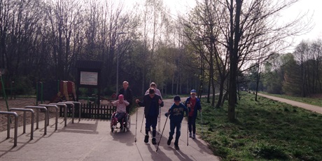 Powiększ grafikę: Pięcioro uczniów i nauczyciel na ścieżce w parku idą z kijkami, nauczyciel prowadzi wózek.