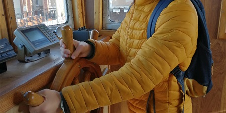 Powiększ grafikę: Chłopiec w żółtej kurtce i okularach stoi w kabinie kutra, trzyma drewniany ster.