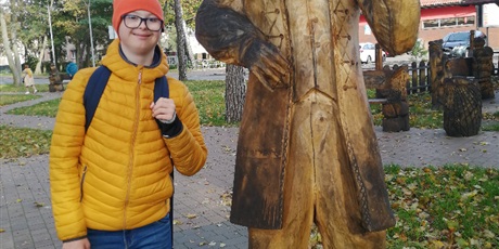 Powiększ grafikę: Chłopiec w żółtej kurtce stoi obok drewnianego posągu przedstawiającego kaszuba. Z tyłu widać drzewa.