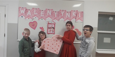 Powiększ grafikę: Dwie dzoiewczynki i dwóch chłopców stoją na tle napisu "Walentynki". Dziewczynki trzymaja razem duży karton w kształcie serca.
