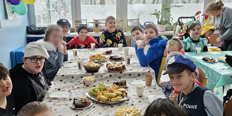 Powiększ grafikę: Uczniowie w strojach karnawałowych siedzą przy stole, na którym stoją owoce i ciastka.