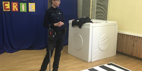 Powiększ grafikę: Pani policjantka stoi w sali gimnastycznej na niebieskim tle z napisem "Bezpieczne ferie", przed nią leży na podłodze makieta przejścia dla pieszych.
