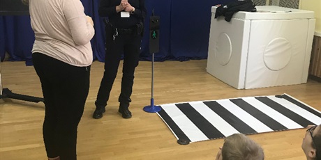 Powiększ grafikę: Dziewczynka stoi obok pani policjantki, na podłodze leży makieta przejścia dla pieszych oraz stoi atrapa sygnalizatora świetlnego.
