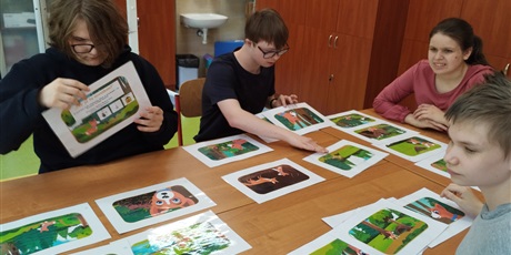 Powiększ grafikę: Uczniowie siedzą dookoła stołu, na którym rozłożone są obrazki.