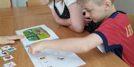 Powiększ grafikę: Chłopiec przykleja PCSy na kartce w książce, obok stoi dziewczynka.