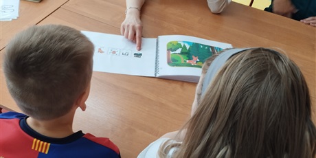 Powiększ grafikę: Chłopiec i dziewczynka siedzą przy stoliku, na którym leży otwarta książka.