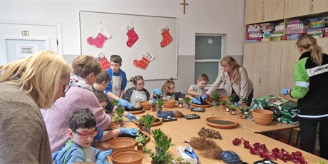 Powiększ grafikę: Uczniowie siedzą dookoła stołu, pfrzed nimi stoją puste doniczki i zielone kwiatki. 