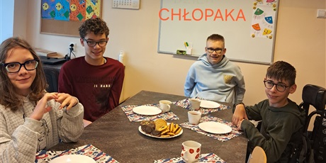 Powiększ grafikę: Czworo uczniów siedzi przy stole, na którym są ciasteczka, talerzyki i kubeczki.