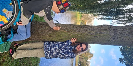 Powiększ grafikę: Dziewczynka i chłopiec na wózku stoją obok drzewa, które dotykają. Za nimi widać staw.