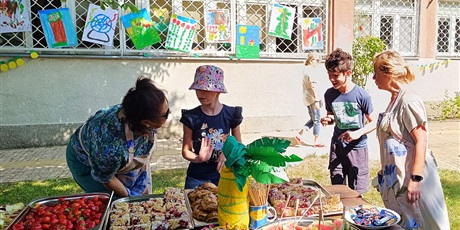 Powiększ grafikę: Dziewczynka i chłopiec oraz dwie nauczycielki stoją przy stole piknikowym pod drzewem, na stole stoją owoce, pokrojone ciasto oraz owocowe szaszłyki. W tle widać prace plastyczne dzieci.