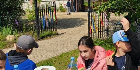 Powiększ grafikę: Uczniowie siedza przy stole piknikowym przed szkoł. Na stole stoją na talerzach truskawki i pokrojony arbuz.