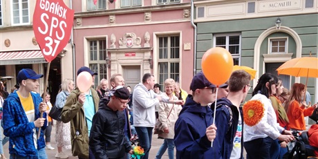 Powiększ grafikę: Uczniowie idą w marszu ulicami Starego Miasta. Jeden uczeń trzyma tarczę z nazwą szkoły, inni trzymają pomarańczowe balony i parasolki.