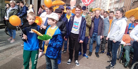 Powiększ grafikę: Uczniowie z nauczycielami idą w marszu ulicami Starego Miasta. Jeden uczeń trzyma tarczę z nazwą szkoły, inni trzymają pomarańczowe balony i parasolki.