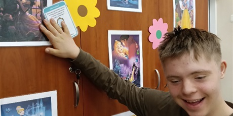 Powiększ grafikę: Chłopiec stoi przy szafie z wizerunkami księżniczek, trzyma kartęz opisem na jednym zdjęciu księżniczki.