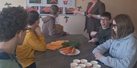 Powiększ grafikę: Uczniowie siedzą przy stole, na którym leżą marchewki i stoją kubeczki. Nauczycielka stoi obok i mówi.