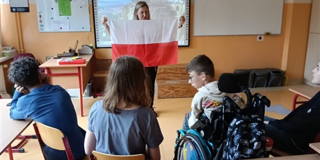 Powiększ grafikę: Uczniowie siedzą w rzędzie, przed nimi stoi pani nauczycielka, która trzyma rozłożoną flagę Polski.