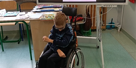 Powiększ grafikę: Chłopiec na wózku stoi na środku klasy. Prezentuje gest, prezentowany na obrazku wyświetlanym na tablicy za nim.