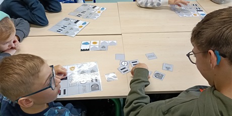 Powiększ grafikę: Uczniowie siedzą dookoła stołu, układają obrazki na planszach.