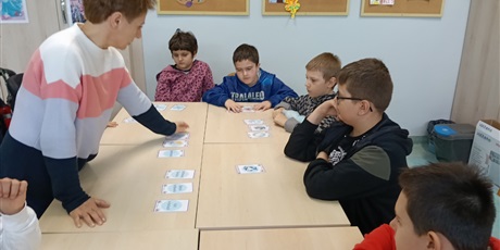 Powiększ grafikę: Uczniowie siedza dookoła stołu, nauczycielka rozkłada na stole karty.