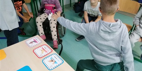 Powiększ grafikę: Uczniowie siedzą na krzesełkach. Chłopiec stoi w pozie pokazanej na obrazku trzymanym przez nauczyciela.