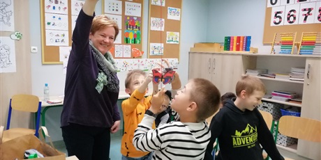 Powiększ grafikę: Uczniowie i nauczyciel stoją na środku klasy. Nauczyciel trzyma na sznurku motylka, uczniowie satarają się go złapać.