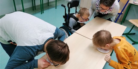 Powiększ grafikę: Troje uczniów liże talerzyki stojące przed nimi na stole. uczniowi na wózku pomaga nauczyciel.