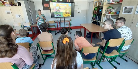 Powiększ grafikę: Uczniowie siedzą na krzesełkach, w tle tablica, na której wyświetlany jest film o pszczołach oraz pani pielęgniarkastoi przy tablicy i coś mówi.