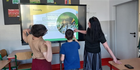 Powiększ grafikę: Dwóch uczniów i nauczycielka stoją przy tablicy, na której wyświetlany jest slajd o pszczołach. 