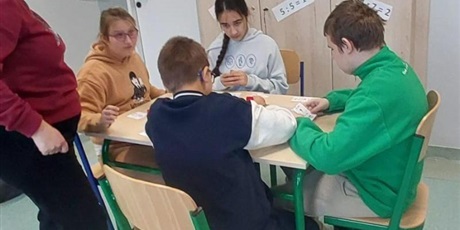 Powiększ grafikę: Uczniowie siedza przy stoliku, obok stoi nauczycielka, liczą.