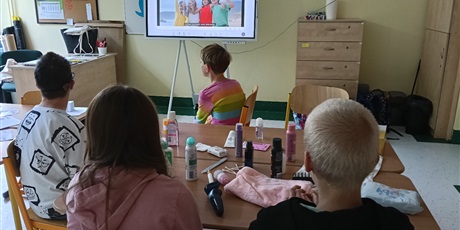 Powiększ grafikę: Uczniowie siedzą przy stoliku, na którym stoją różne kosmetyki.