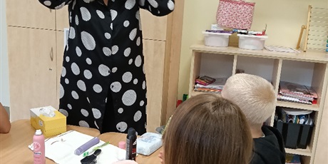 Powiększ grafikę: Chłopiec i dziewczynka siedzą przy stoliku, na którym stoją różne kosmetyki. Obok stolika stoi nauczycielka, trzyma się za pochyloną głowę.