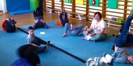 Powiększ grafikę: Uczniowie w sali gimnastycznej leżą w podniesionymi do góry nogami na macie. Nauczycielka siedzi po turecku, obserwuje dzieci.