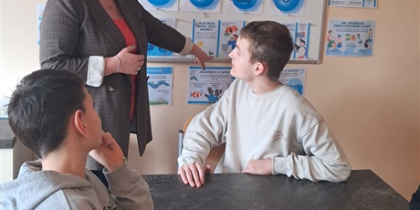 Powiększ grafikę: Dwóch chłpoców siedzi przy stoliku, nauczycielka pokazuje karty i mówi.