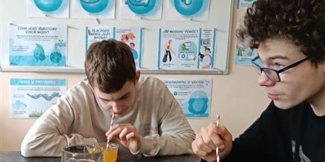 Powiększ grafikę: Uczniowie dmuchają przez słomkę do pomarańczowej cieczy w szklance.