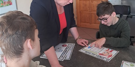 Powiększ grafikę: Dwóch chłopców siedzi przy stole, obok stoi nauczycielka, chłopiec pokazuje element na obrazku.