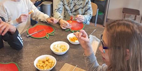 Powiększ grafikę: Uczniowie siedzą przy stole, przed każdym leży deska do krojenia, na środku stołu w miseczkach leżą pokrojone owoce. Uczniowie robią owocowe szaszłyki.