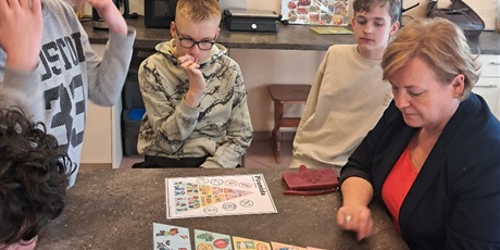 Powiększ grafikę: Uczniowie i nauczycielka siedzą przy stole, na którym leży ułożona piramida żywieniowa.