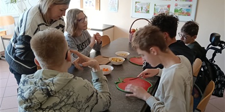 Powiększ grafikę: Uczniowie siedzą przy stole, kroją na dekach owoce w kostkę.
