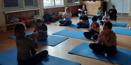 Powiększ grafikę: Uczniowie siedzą na matach w sali gimnastycznej, wykonują ćwiczenia prezentowane przez nauczycielkę.