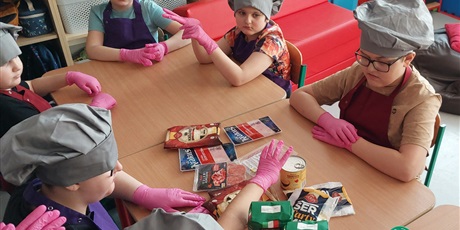 Powiększ grafikę: Uczniowie siedzą dookoła stołu, na którym leżą produkty do pizzy. Wszyscy mają na głowie szarą czapkę kucharską.