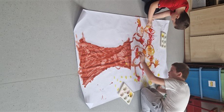 Powiększ grafikę: Na podłodze leży namalowane na dużym arkuszu papieru drzewo, dwóch chłopców odbija swoje pomalowane na kolorowo dłonie.