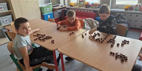 Powiększ grafikę: Trzech chłopców siedzi przy stoliku, przed każdym z nich leżą kasztany.