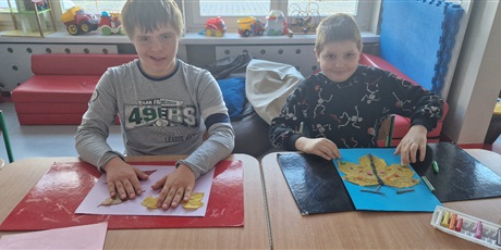 Powiększ grafikę: Dwóch chłopców siedzi przy stoliku, przed nimi przed nimi leżą kartki z przyklejonymi dużymi liśćmi klonu.