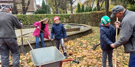 Powiększ grafikę: Dwóch chłopców i dziewczynka grabią liście na trawniku. Obok stoi taczka, dwóch panów ogrodników pomaga dzieciom.