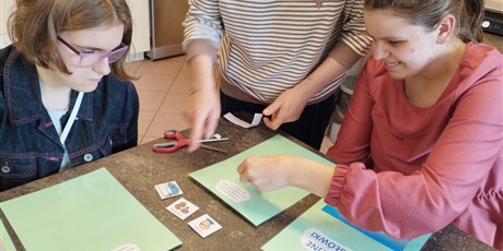 Powiększ grafikę: Dwie dziewczynki siedzą przy stole przed nimi leżą zielone kartki i symbole PCS. Obok stoi kobieta, która pokazuje symbol palcem.