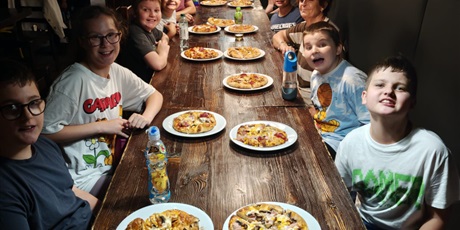 Powiększ grafikę: Dzieci siedzą przy długim stole. Przed każdym stoi talerz z samodzielnie wykonaną pizzą.